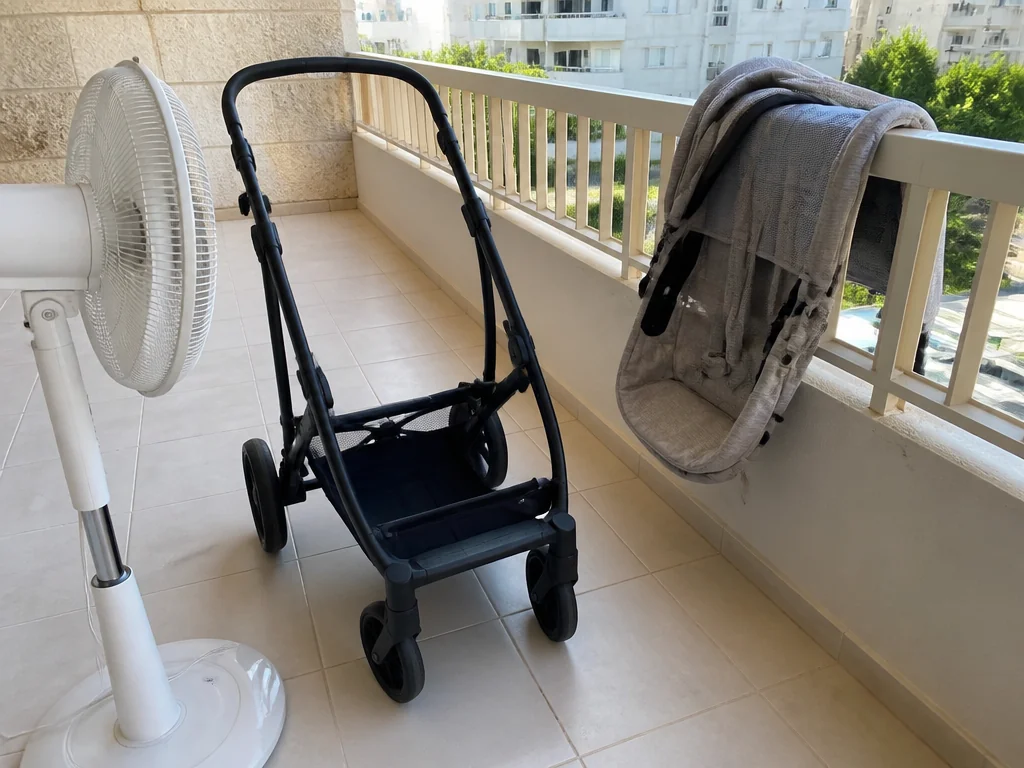 עגלת תינוק פתוחה במרפסת מוצלת עם מאוורר עומד מכוון אליה לייבוש